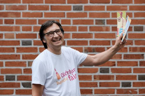 Mann in T-Shirt mit dem Logo von Oldenburg handelt fair und Oldenburg Fair Flyer in den Händen. Foto: © J. Bädeker