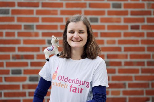 Eine Frau, Julia Ingensiep, steht vor einer roten Backsteinwand mit einem Kuscheltier-Alpaka auf der Schulter und trägt ein T-Shirt mit der Aufschrift "Oldenburg handelt fair. Foto: © J. Bädeker